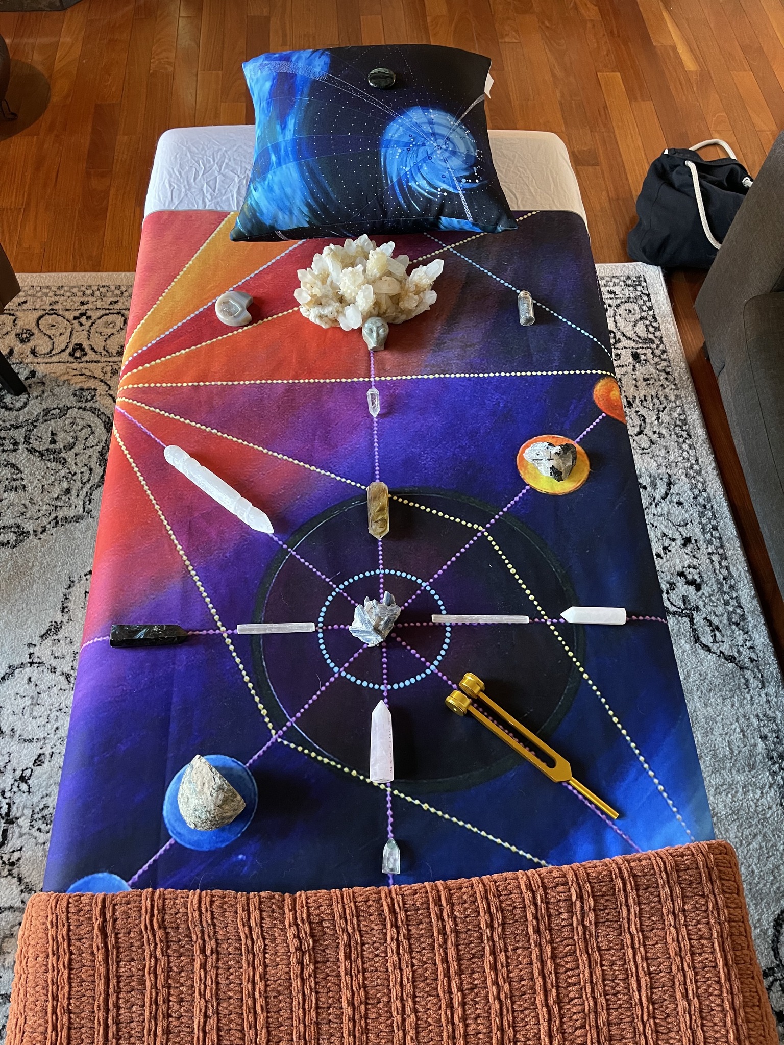 Massage table with a colourful tapestry called Orbital Crown draped over the table, crystals placed in certain grid lines for energy and healing. A colourful pillow with artwork called 